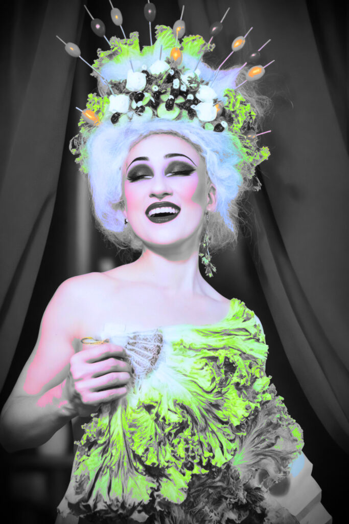 Artiste burlesque Lady Vergeture souriante, vêtue d'une robe évoquant une salade, avec une coiffe ornée de légumes comme des tomates, des olives et des feuilles de salade.